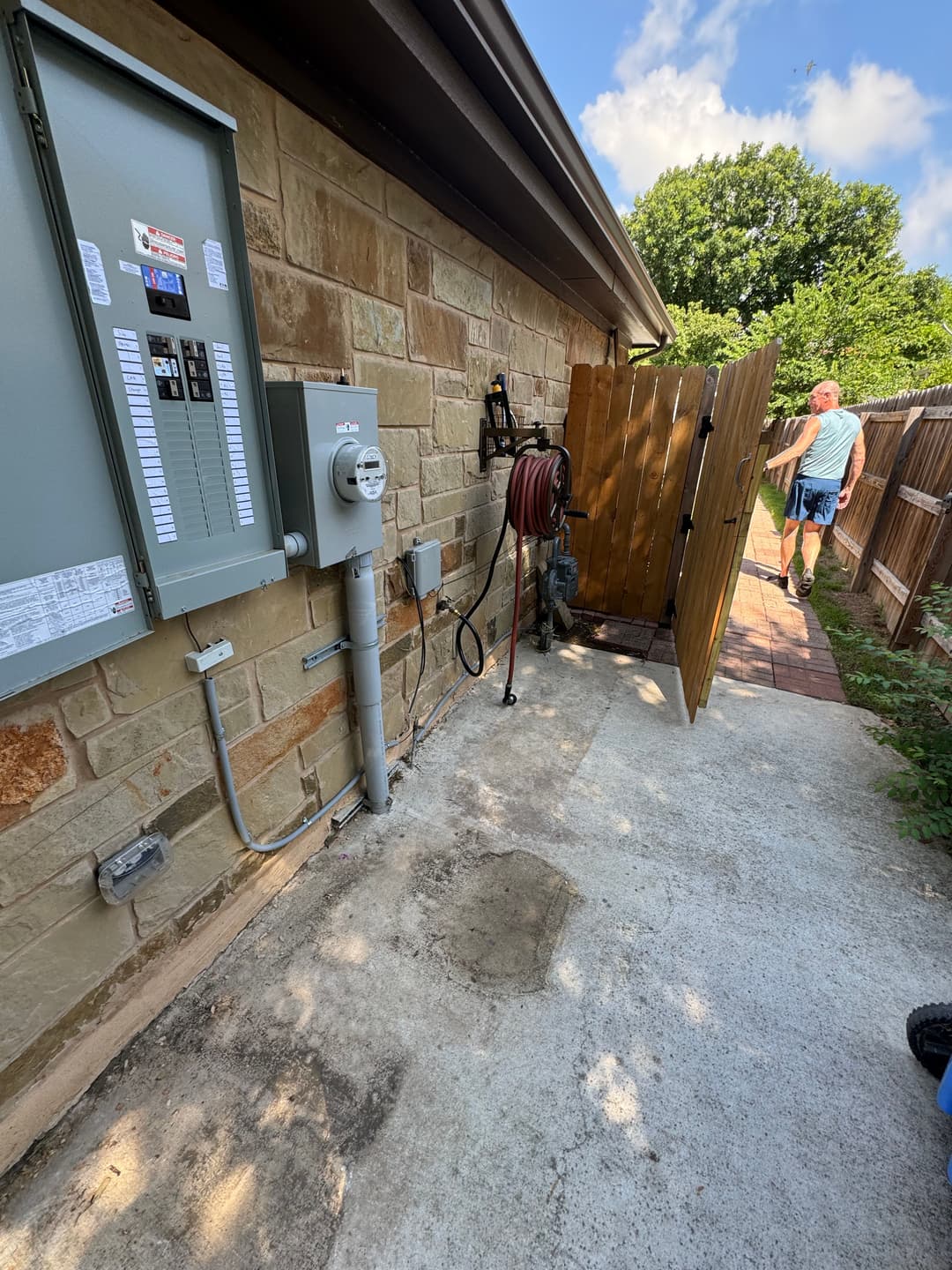 Electrical panel and utility meter on a stone wall beside a wooden gate and hose reel.