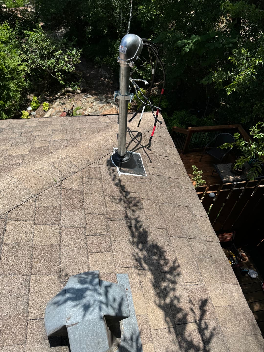 Aerial view of a roof with a metal antenna and shadow on shingles surrounded by greenery.