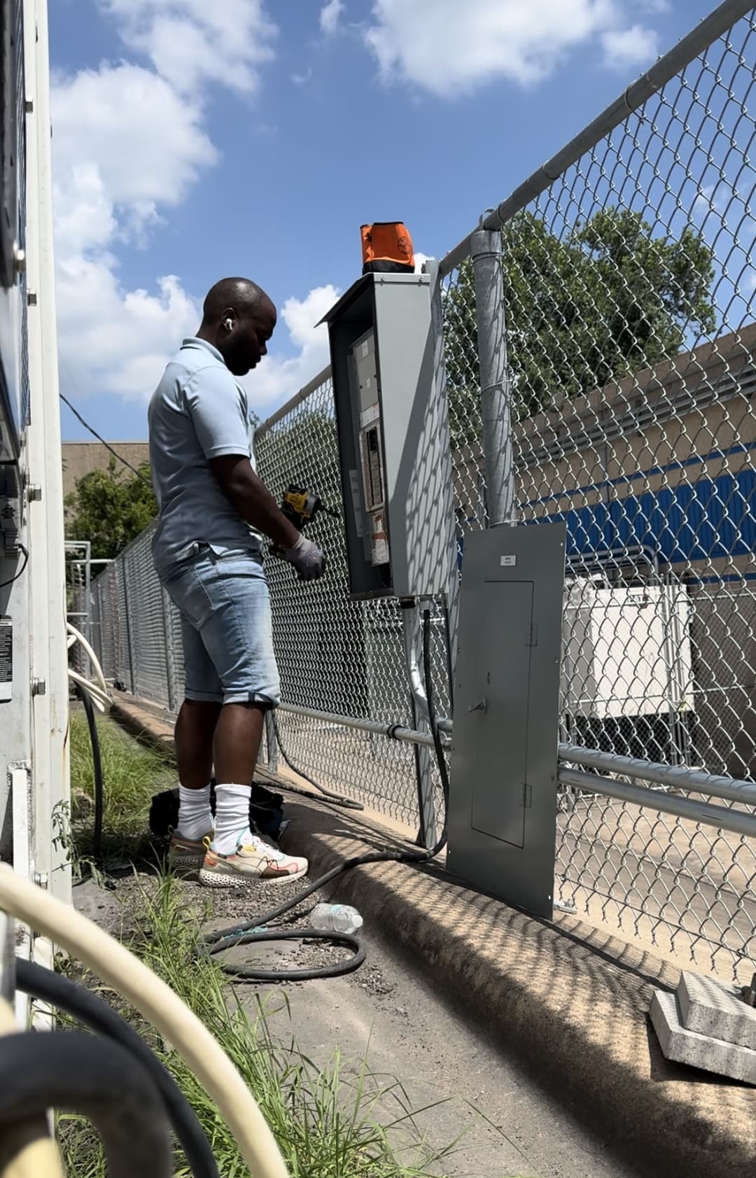 Man in denim shorts using a service panel outside a fenced area on a sunny day.