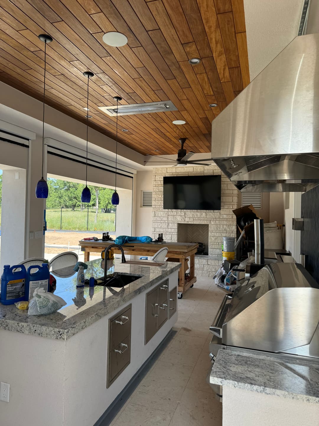Modern outdoor kitchen with stainless steel appliances, stone fireplace, and wooden ceiling.