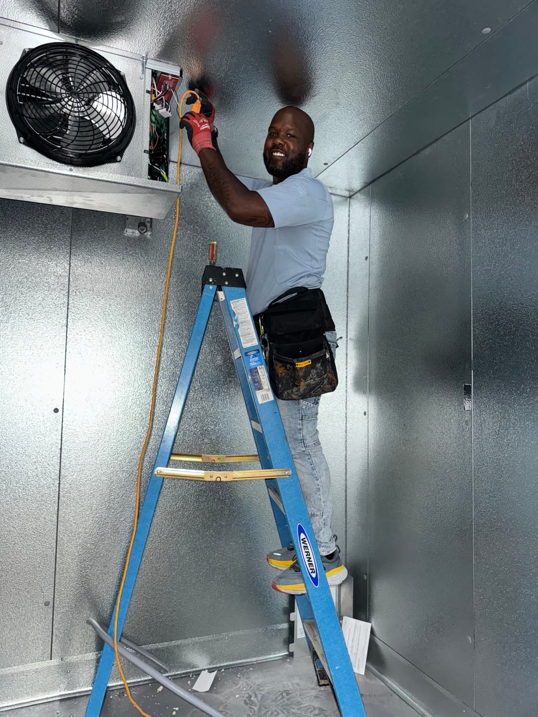 Electrician on ladder working on wiring inside a metallic room with ventilation fan.