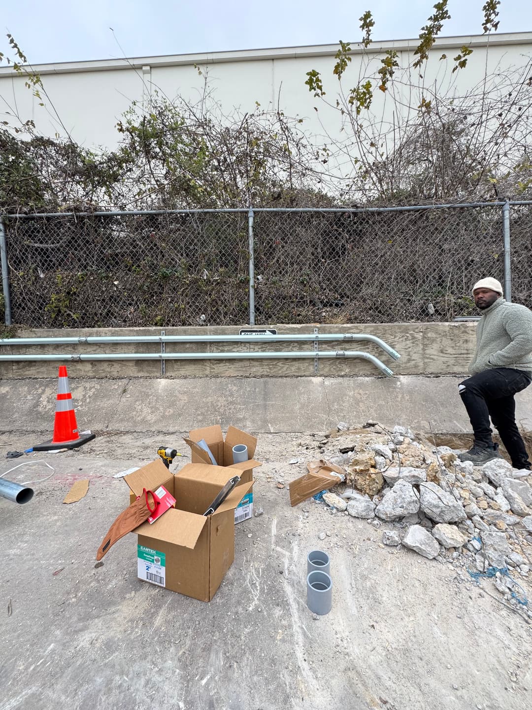 Construction site with debris, pipes, and a worker near a fenced area. Safety cones visible.