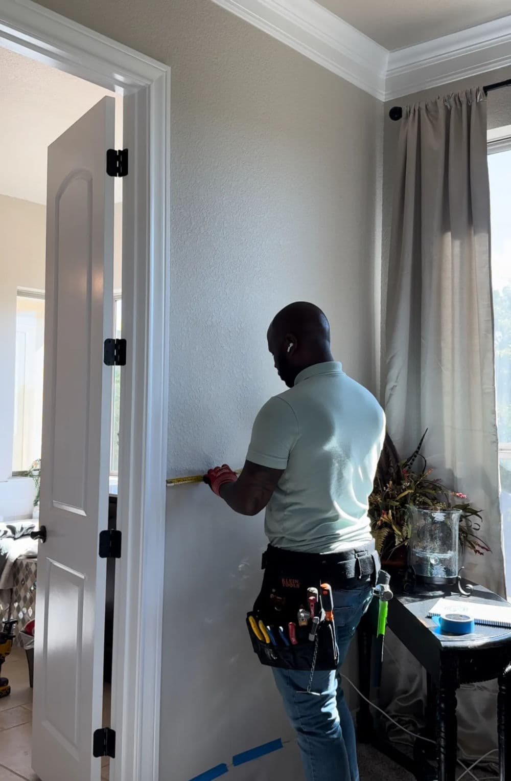 Man painting a wall with tools in a tool belt, wearing a light blue shirt in a well-lit room.