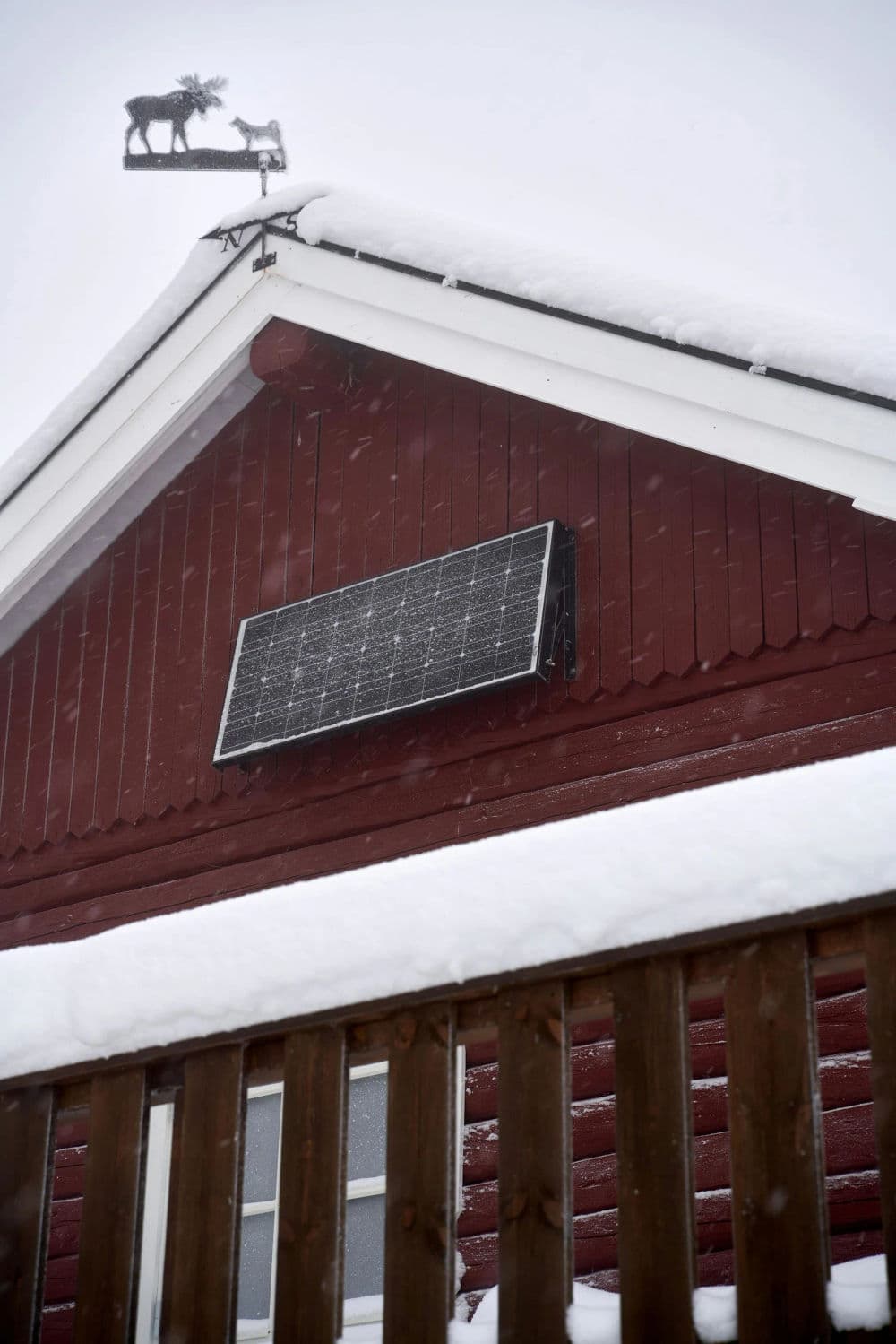 Solar panel installed on a red cabin roof, with snow covering the eaves and a weather vane.
